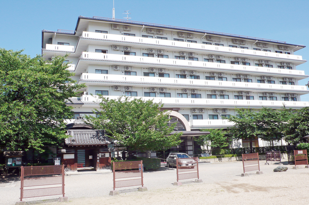 介護施設の外観。白色5階建てビルで、複数のエアコン室外機と балконが見える。玄関周辺に木立がある。